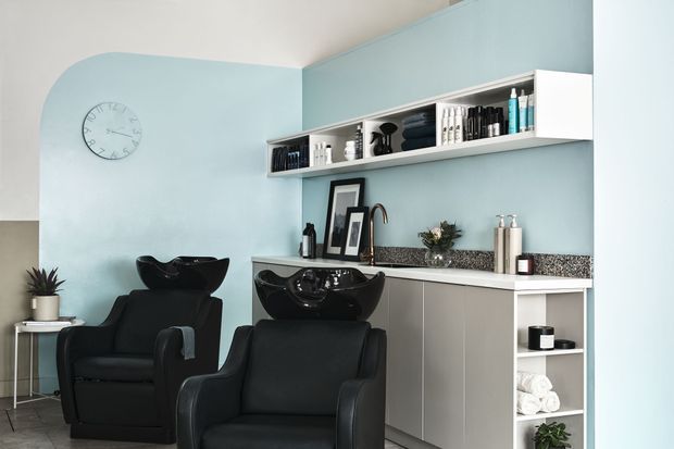 A compact, contemporary hair salon wash area with two black shampoo stations and reclining salon chairs, set against a pale blue wall. White built-in cabinetry and open shelving provide storage for hair products, towels, and accessories, with a speckled stone-look benchtop and decorative styling items. The room has a clean, professional, softly coordinated appearance.