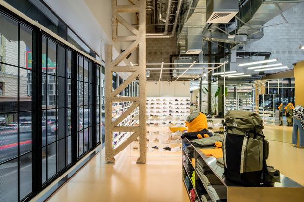 A contemporary retail showroom interior with exposed concrete columns, services, ductwork and ceiling structure. Full-height black-framed glazing lines the left side, while illuminated wall displays of footwear and central product tables showcase outdoor clothing and equipment. The space has polished pale flooring, bright linear lighting and an industrial, open-plan fit-out.
