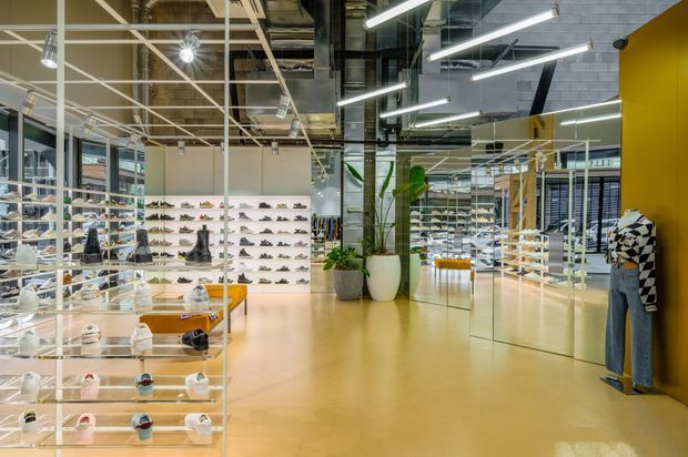 Interior view of a contemporary retail footwear and fashion showroom, with extensive wall-mounted and freestanding shoe displays, glass shelving, clothing racks, full-height mirrors, indoor plants, polished pale flooring, and exposed industrial-style ceiling services with linear lighting.