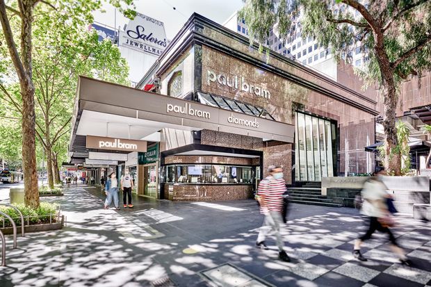 Street-level view of a multi-storey urban jewellery retail storefront, with prominent Paul Bram diamond signage, dark polished stone-clad facade, projecting awnings, large display windows, broad entry steps, and a tree-lined pedestrian streetscape.