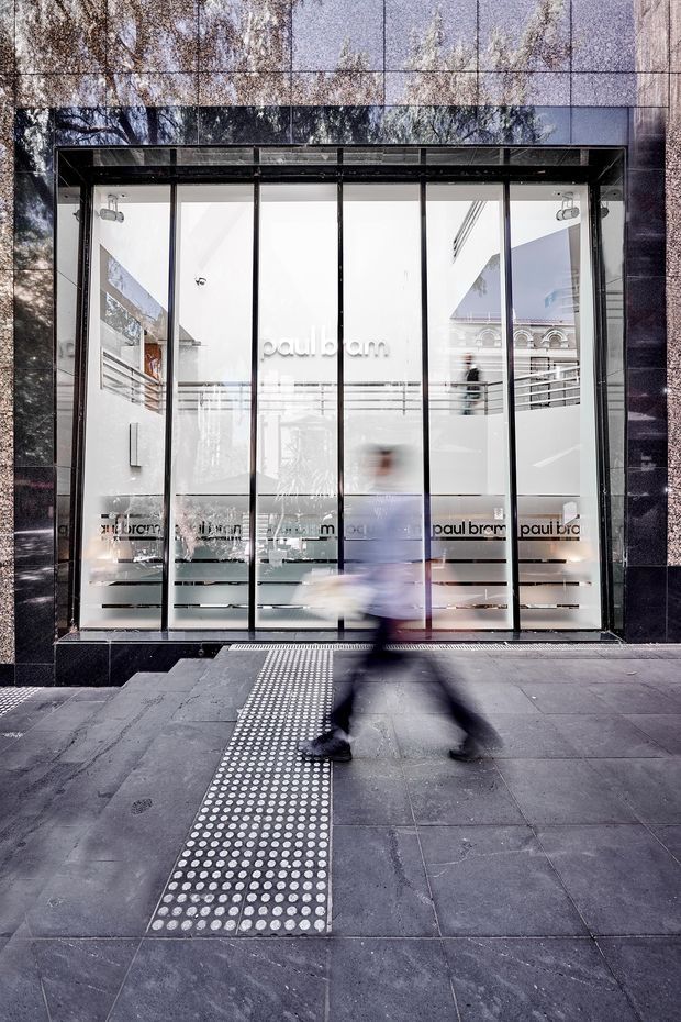Street-level photograph of a contemporary retail storefront with a highly glazed black-framed facade. The bright white commercial interior is visible through the floor-to-ceiling glass, with Paul Bram branding, shelving, and a mezzanine or upper-level balcony. Dark stone or tiled cladding frames the entrance, and a tactile guidance strip runs across the dark paved footpath. A blurred pedestrian passes in the foreground.