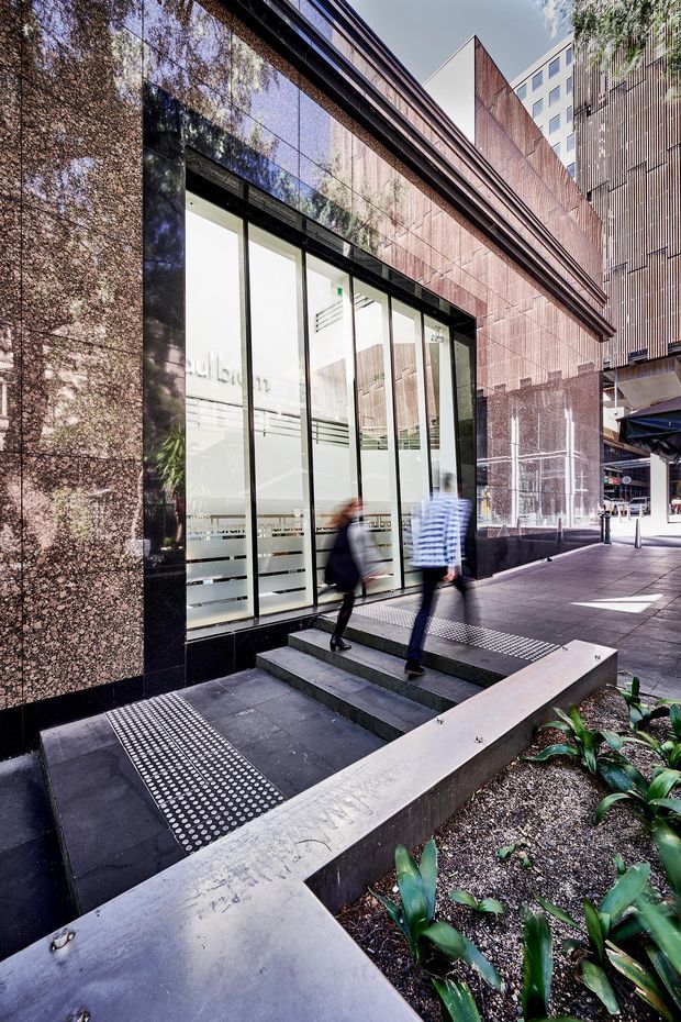 Street-level exterior view of a contemporary commercial building entrance. The facade is clad in dark polished stone or stone-look panels with vertical ribbed metal detailing, large glazed doors and windows, and a stepped accessible entry with tactile paving. A landscaped planter and urban pedestrian walkway occupy the foreground.