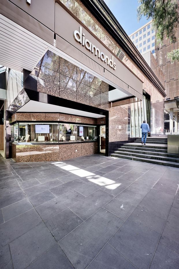 Street-level view of the Diamond jewellery store entrance in an urban commercial setting. The facade features polished reddish-brown stone or granite panels, dark trim, extensive glazing, a projecting canopy, illuminated retail display windows, and broad dark tiled paving with steps leading to the entrance. Tall surrounding office buildings and street trees are visible.