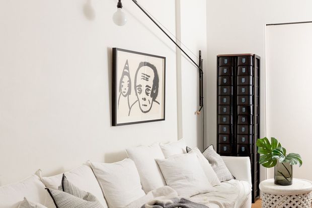 Bright, compact living room with a white upholstered sofa layered with neutral cushions and throws. The room features a framed monochrome artwork, a tall dark wood storage cabinet, exposed black wall-mounted lighting cables, a potted tropical plant, and a small round side table. The overall look is clean, eclectic, and lightly industrial with strong neutral tones.