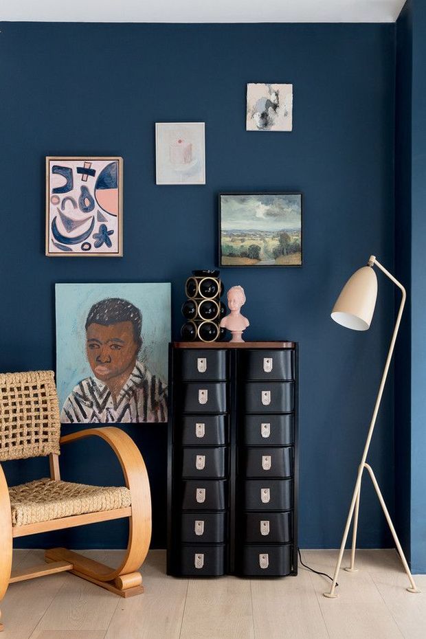 A stylish interior corner with a deep navy-blue painted wall, pale timber flooring, and a black vintage-style drawer chest. The room features a woven bentwood armchair, a sculptural floor lamp, framed artwork, a portrait leaning against the wall, a small bust, and decorative storage. The composition has a curated, artistic and retro character with strong contrast between the dark wall and light furnishings.