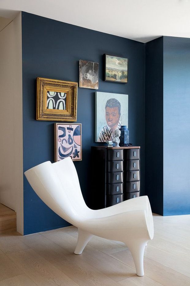 A stylish interior corner with deep navy-blue walls, pale timber flooring, and a sculptural white lounge chair in the foreground. A dark wood chest of drawers stands against the feature wall, topped with decorative objects and surrounded by an eclectic arrangement of framed artwork. The space is bright with a refined, gallery-like atmosphere.