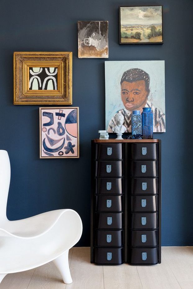 A styled interior vignette with a deep navy-blue wall, pale timber flooring, a tall black drawer unit, and a white sculptural lounge chair. Several framed artworks are arranged above and around the cabinet, with blue decorative vessels and small objects displayed on top. The composition has a curated, art-focused look with strong contrast between the dark wall and light furniture.