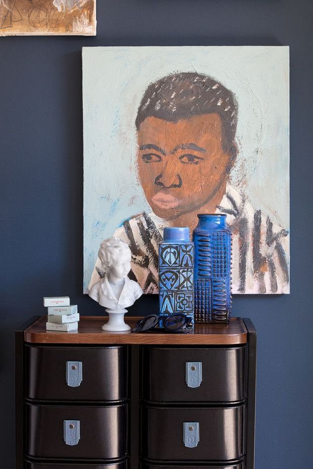 A styled interior vignette featuring a dark navy-blue wall, a large expressive portrait painting, and a vintage dark timber or lacquered chest of drawers with metal pulls. The console is accessorised with a white classical bust, stacked books, blue decorative ceramic vessels, and glasses. The composition has a curated, eclectic and art-focused character with strong dark-to-light contrast.