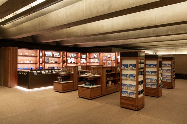 A warmly lit retail showroom or visitor shop with extensive dark timber shelving, display cabinets, product racks, and a central checkout or display counter. The space has a low, exposed concrete ceiling with dark structural beams, polished neutral flooring, and a museum-like commercial interior arrangement.