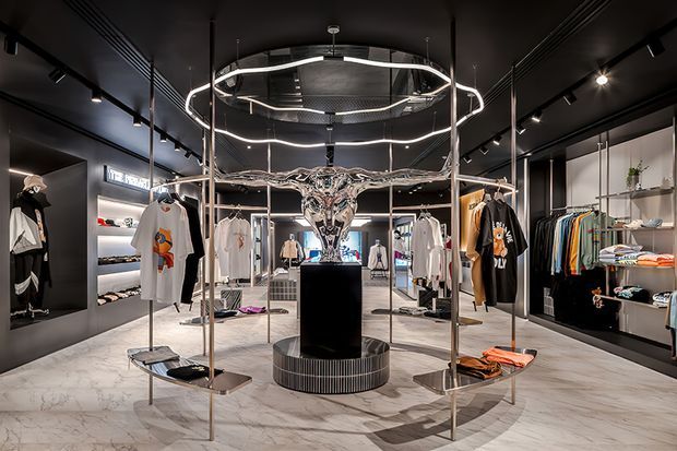 A high-end contemporary fashion retail showroom with a dark, dramatic interior. Clothing is displayed on freestanding metal racks and wall-mounted shelving around a central sculptural metallic bull head mounted above a black pedestal. Large circular and linear ring lights create a strong focal point, while pale marble-look flooring contrasts with the black ceiling, walls, cabinetry, and fixtures.