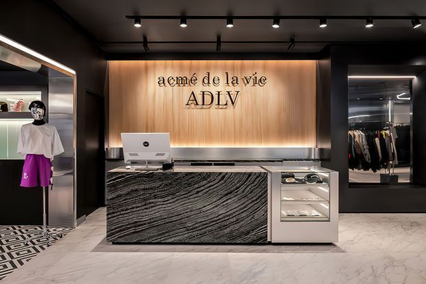 A sophisticated fashion retail boutique interior viewed toward a central reception and checkout counter. The space features black walls and ceiling, track lighting, a warm vertical timber feature wall with the 'acmé de la vie ADLV' logo, a dramatic dark veined stone-clad counter, white display cabinetry, marble-look flooring, clothing racks, and a mannequin display. The overall design is high-contrast, polished, and luxurious.