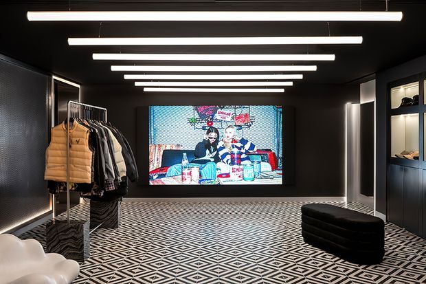 A dramatic, high-contrast retail showroom or fashion display space with black walls and ceiling, linear suspended LED lighting, a large colourful digital artwork or screen, patterned geometric flooring, a clothing rack, illuminated display shelving, and a black upholstered bench.