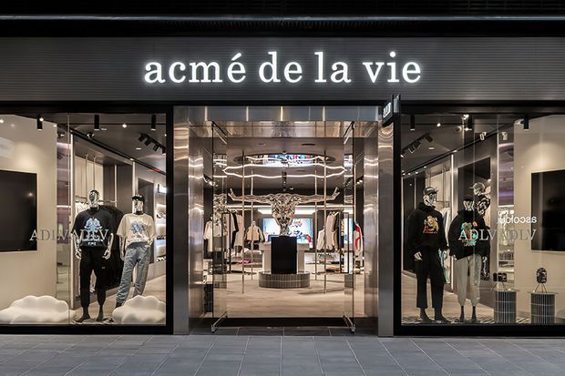 Front-facing view of a contemporary fashion retail storefront with a black facade, illuminated “acmé de la vie” signage, expansive display glazing, mannequins, and a central glass entrance. The interior features dark ceilings, bright display lighting, reflective metallic framing, and clothing displays.