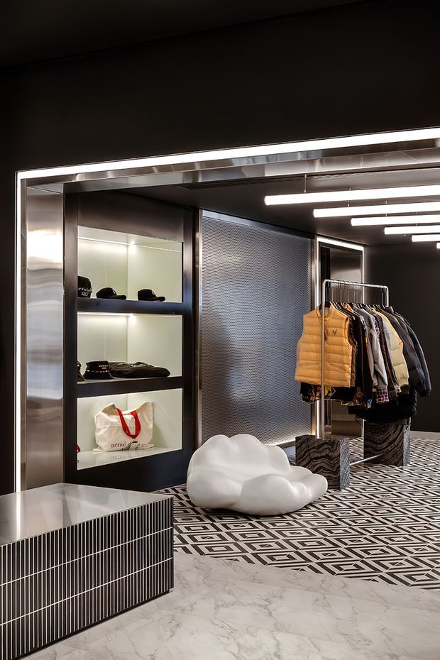 A dramatic contemporary retail interior with dark walls and ceilings, illuminated recessed display shelving, stainless-steel framing, patterned geometric flooring, and a marble-look foreground floor. Clothing is displayed on a freestanding rack, while sculptural white seating and striped built-in cabinetry add strong decorative elements. The space has a high-contrast monochrome palette with metallic accents and gallery-like lighting.