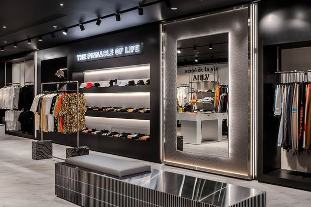 A sophisticated contemporary clothing retail interior with black walls and ceiling, integrated illuminated shelving displaying hats and accessories, hanging garment racks, and a large decorative mirror framed by textured metallic trim. A long central display bench with a reflective metal surface and upholstered cushion anchors the space. Track lighting and concealed shelf lighting create a dramatic, high-contrast boutique atmosphere.