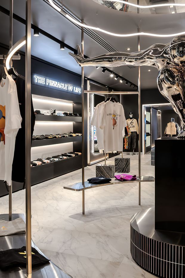 A luxurious contemporary clothing retail interior with dark display walls, illuminated shelving, marble-look flooring, slim metallic clothing racks, sculptural chrome artwork, and curved integrated LED lighting. Apparel and accessories are displayed throughout the polished showroom.