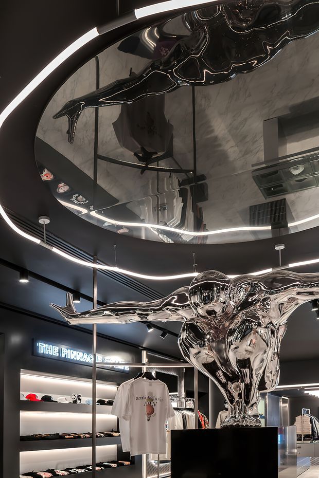 A dramatic contemporary retail interior with a predominantly black ceiling, curved linear LED lighting, illuminated merchandise shelving, clothing displays, and a large highly reflective chrome sculptural figure suspended or mounted above a black pedestal. A mirrored ceiling feature reflects the sculpture and interior below, creating a futuristic, glossy atmosphere.