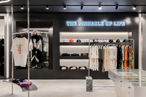 A sleek contemporary clothing retail interior with a predominantly black and grey fit-out. The space features illuminated wall shelving, hanging garments, display mannequins, glass-topped merchandise counters, track lighting, mirrored panels, and a neon sign reading “THE PINNACLE OF LIFE.”