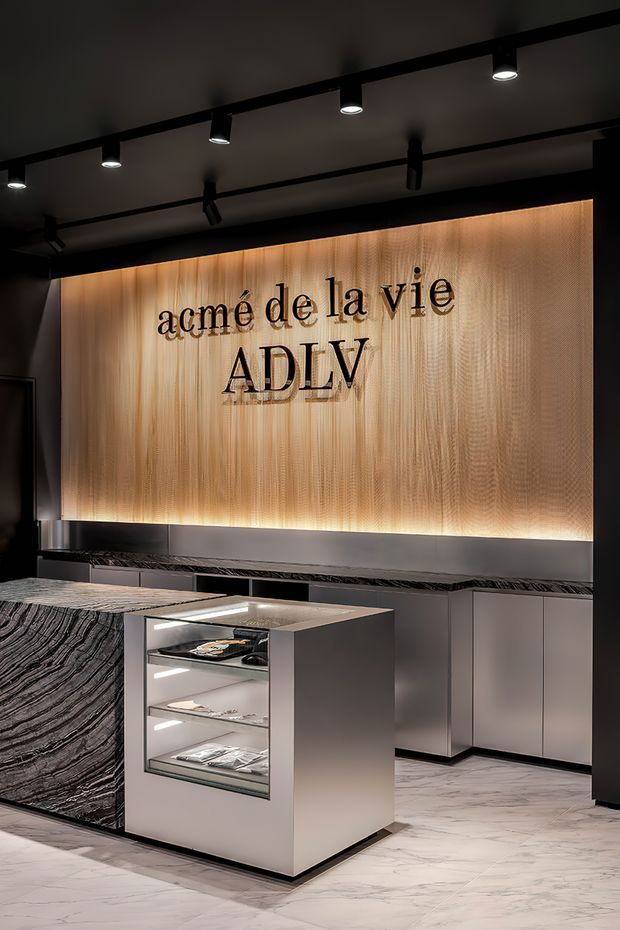 A dark, refined commercial interior featuring a branded reception or retail display area. A large backlit light-wood feature wall carries the “acmé de la vie ADLV” lettering, with black track lighting above, dark cabinetry, a dramatic veined stone counter, and a central illuminated glass display island. The space has a luxurious, contemporary showroom aesthetic.