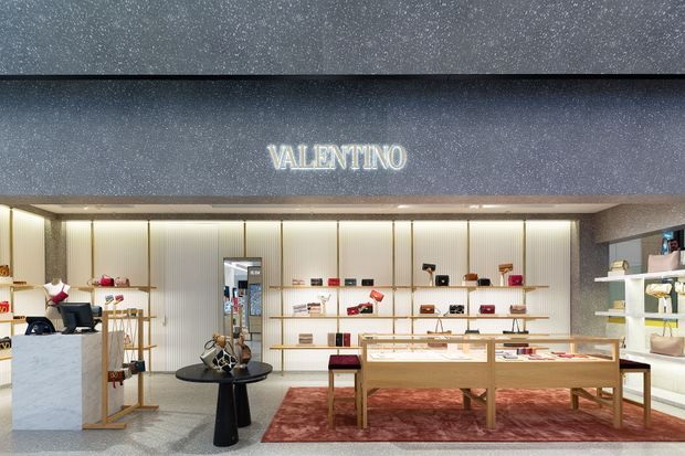 A refined Valentino retail boutique interior with illuminated ribbed wall panels, warm integrated lighting, glass and timber display cases, pale stone counters, and a deep red area rug. Luxury handbags and accessories are arranged throughout the shelving and display tables, with the Valentino logo prominently featured above the rear wall.