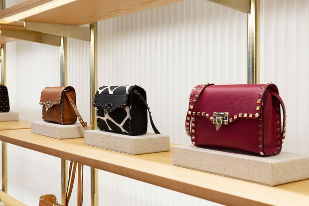 Close-up retail display of several handbags arranged on light timber shelves, each placed on rectangular upholstered plinths. The backdrop consists of white vertically ribbed translucent panels, with warm integrated lighting and timber framing creating a refined boutique showroom presentation.