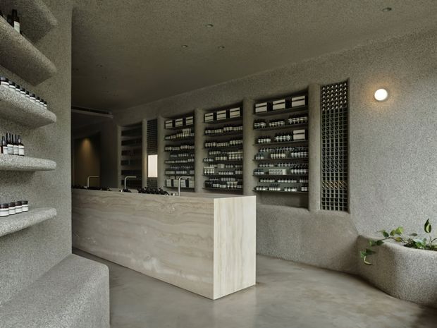 A minimalist retail or commercial interior, likely a beauty or skincare shop, with a pale stone or concrete reception counter, integrated wall shelving filled with products, textured grey plaster walls, terrazzo-like built-in seating, and a perforated breeze-block screen. The space has a muted monochromatic palette, concealed or recessed lighting, and sparse greenery.