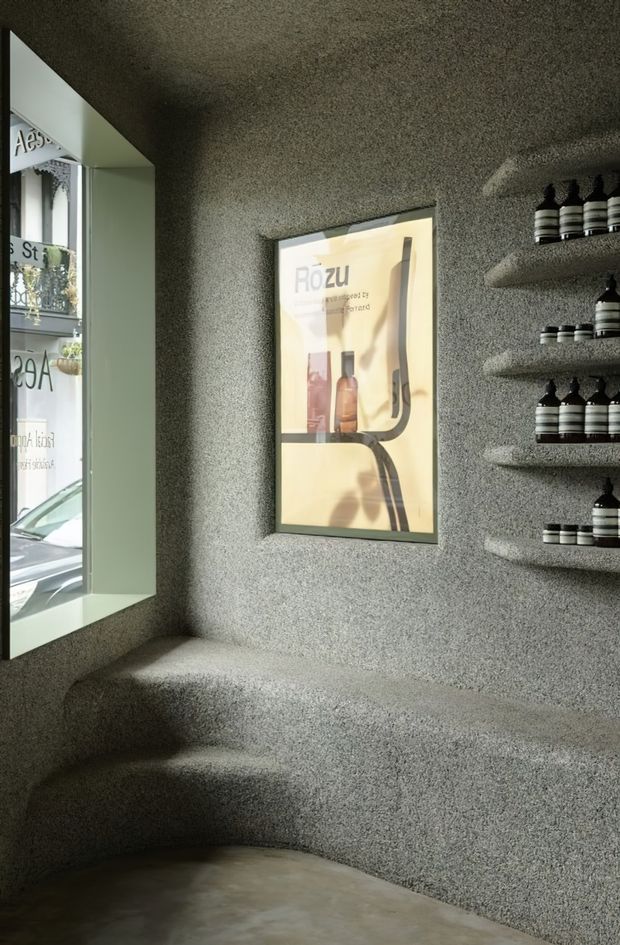 A compact retail or commercial interior with a highly textural grey sprayed or pebble-finished wall, integrated curved bench seating, recessed display shelves holding small black-and-white product bottles, and a framed illuminated advertisement. A large angled window overlooks a street and provides natural light. The space has a sculptural, monochromatic, minimalist fit-out.