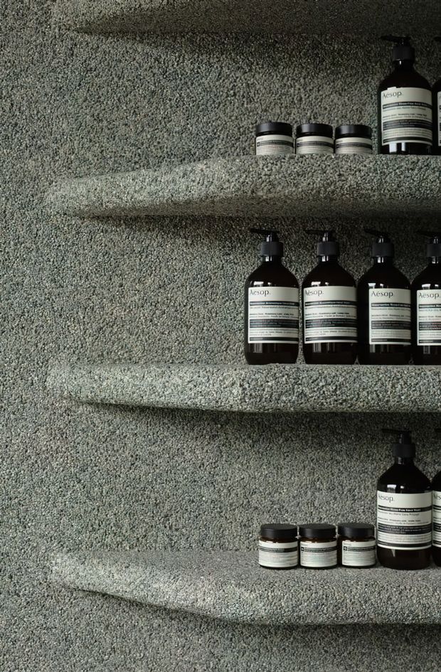 Close-up of dark Aesop skincare bottles and small jars arranged on thick, rounded built-in shelves. The shelves and surrounding wall are covered in a heavily textured grey-green finish, creating a monochromatic, tactile retail display.