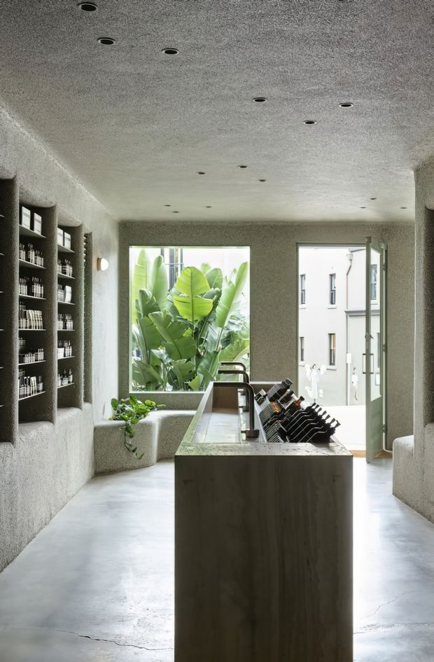 A narrow, refined wine shop or tasting space with a central stone or concrete island displaying wine bottles, recessed ceiling spotlights, built-in wall shelving filled with bottles, textured plaster walls, polished concrete flooring, and large openings overlooking lush tropical foliage and an adjacent courtyard building.