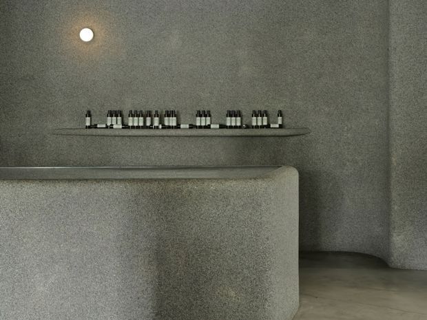 A minimalist grey textured interior with a rounded built-in counter or reception desk in the foreground. A slim dark floating shelf displays small bottles, while a recessed circular ceiling light provides a subtle warm glow. The space has softly curved walls, seamless finishes, and a polished concrete-like floor.