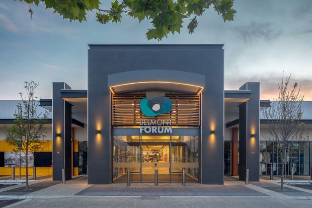 Front-facing exterior view of a contemporary commercial building entrance, with a strong symmetrical facade, dark charcoal cladding, broad entry canopy with integrated lighting, timber-look horizontal screening, glazed double-height doors, and prominent “Belmont Forum” signage. The building is set along a paved pedestrian frontage with young trees and illuminated architectural details.