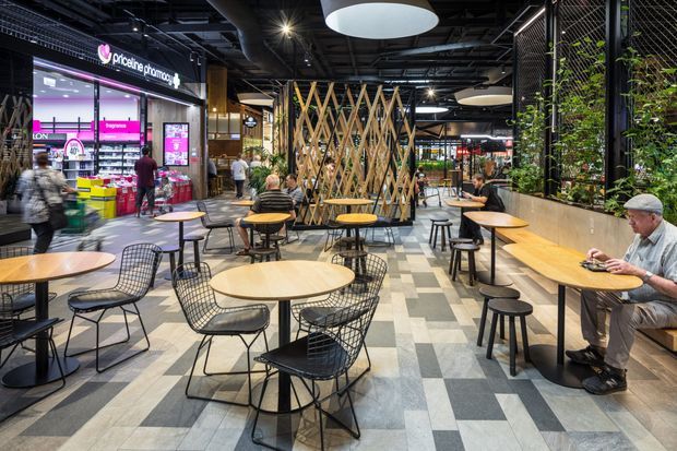 A contemporary indoor shopping-centre dining and seating area with numerous round timber tables, black wire-frame chairs, stools, and built-in bench seating. A retail storefront is visible on the left, while a timber lattice divider and planted metal-framed screen create separation through the centre and right side. The space has an exposed dark ceiling with track and pendant lighting, patterned grey tiled flooring, and an informal food-court or communal hospitality layout.