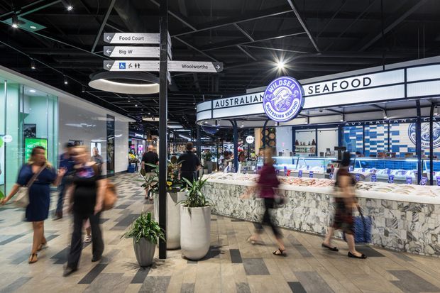 Interior view of a contemporary shopping centre food court, focused on a seafood market-style retail counter with prominent signage, tiled counters, directional wayfinding signs, patterned flooring, indoor planting, and a dark exposed services ceiling. Shoppers are moving through the space, with motion blur suggesting an active commercial environment.