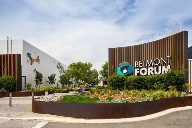Exterior view of the Belmont Forum commercial shopping and entertainment complex. The image shows a landscaped entry plaza with raised rust-toned planter beds, ornamental planting, bollards and paved pedestrian paths. The main facade features a large curved wall with dark vertical slatted cladding, prominent Belmont Forum signage, and adjacent white contemporary building volumes with colorful geometric wall artwork.