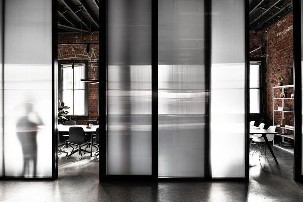 Interior view of a contemporary office or commercial workspace divided by tall translucent sliding or pivoting partitions in black frames. The partitions create semi-private meeting and work areas, with exposed red-brick walls, exposed ceiling structure and services, polished concrete flooring, desks, office chairs, shelving, and pendant lighting visible beyond. The composition is monochromatic and architectural, with strong vertical lines and filtered daylight.