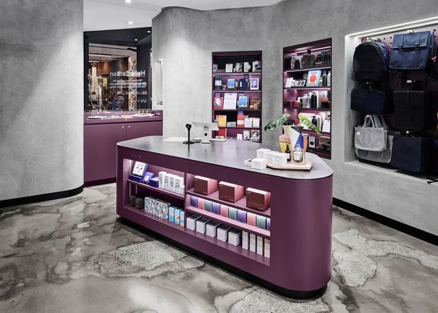 A contemporary retail showroom with a curved plum-coloured display counter and integrated shelving stocked with boxed products and accessories. Dark inset wall shelving, a wall-mounted display of bags, polished concrete-look flooring, and grey textured walls create a refined, boutique commercial interior.