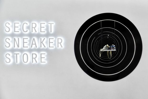 A minimalist commercial retail interior wall featuring a glowing white “SECRET SNEAKER STORE” sign beside a large black circular display with concentric illuminated rings and sneakers suspended at its centre.