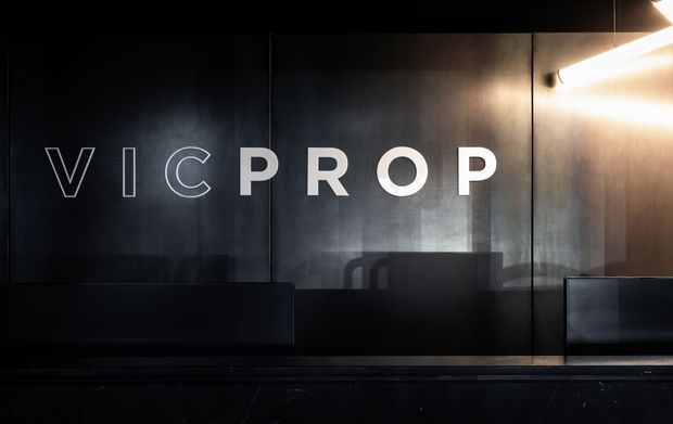 Dark, moody commercial interior featuring a large black or charcoal wall with the illuminated “VICPROP” logo, reflective dark panels, low built-in seating or counters, and a strong linear wall light at the upper right.