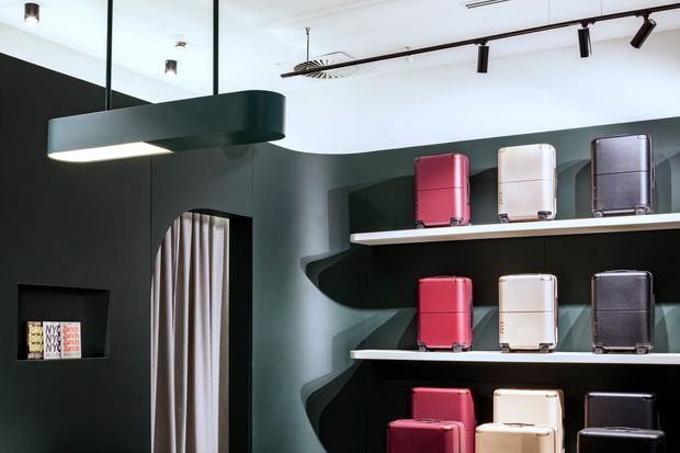 A contemporary retail display interior featuring rows of colourful hard-shell luggage arranged on white floating shelves against a dark sculptural curved wall. The space has a white ceiling with black track lighting, a large black pendant light, a mirrored upper wall, and a curtained arched opening.