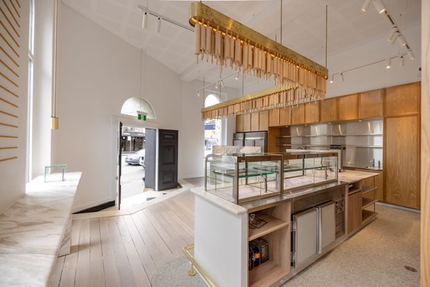 Bright contemporary retail food-service interior with a long central glass display counter, integrated stainless-steel refrigeration and preparation equipment, and extensive warm timber cabinetry. The space has white walls, pale timber flooring, high ceilings, track lighting, large front windows, and distinctive linear brass pendant fixtures with hanging rods. An entrance door opens to the street, reinforcing the commercial shop or café setting.
