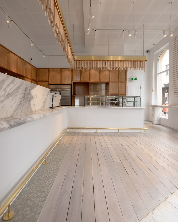 Bright, refined commercial café or bakery interior with a long white service counter, glass display case, timber wall cabinetry, marble-look stone counter and backsplash, pale painted timber floorboards, and brass rail detailing. A large decorative rectangular pendant with hanging elements is suspended over the counter, while track lighting and arched glazing add architectural character.