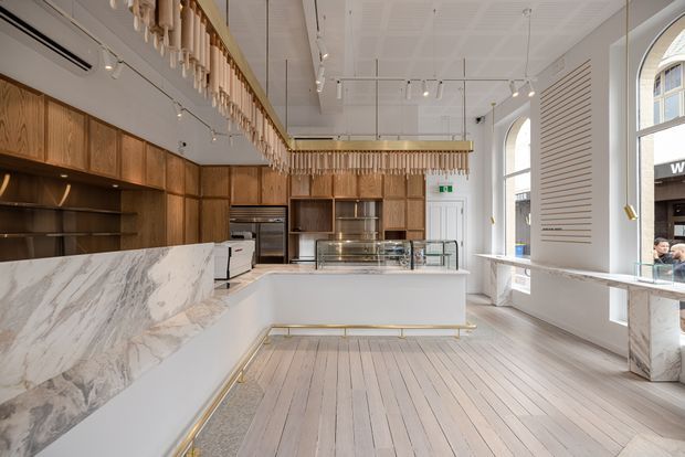Bright, refined commercial food-service interior with a long marble-clad counter, central glass display island, warm timber cabinetry and shelving, white decorative ceiling, brass-toned trim, and tall arched windows. The space has a contemporary yet classic retail-cafe fit-out with carefully integrated display lighting.