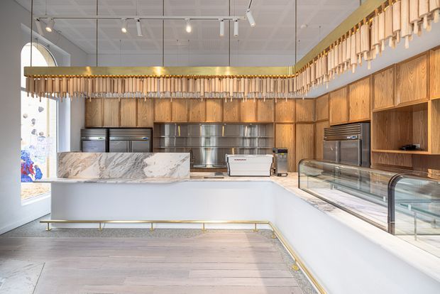 Bright contemporary commercial food-service interior with a large white service counter, marble-look stone counter insert, extensive warm timber cabinetry, stainless-steel kitchen equipment, glass display cases, and a decorative timber pendant installation. Pale timber flooring and a high gridded ceiling create an airy, refined atmosphere.