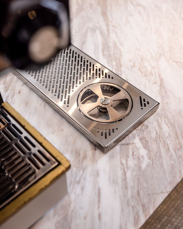 Close-up product-style photograph of a stainless steel rectangular kitchen sink drainboard and strainer assembly set into a pale marble-look benchtop. The drainboard has elongated perforations and a circular removable strainer cover, with a second dark metal drain component partially visible at the lower left.