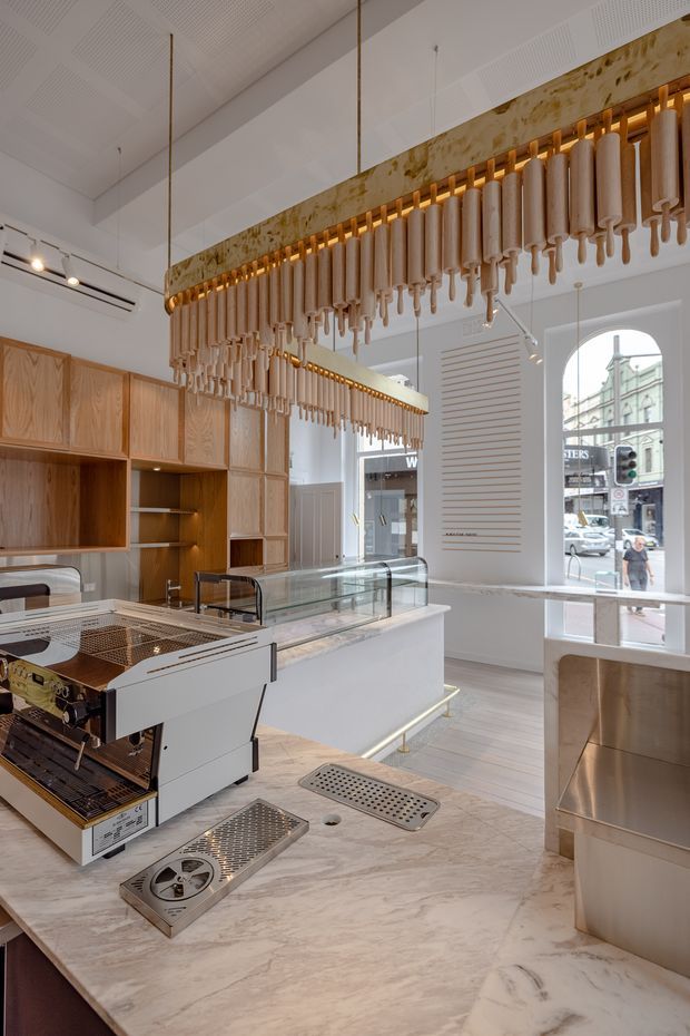 A refined commercial coffee shop interior with a white and pale timber fit-out. The space features a long marble service counter with integrated coffee equipment, glass pastry display case, timber wall cabinetry and shelving, and a striking suspended decorative installation made from rows of light cylindrical elements with warm accent lighting. Tall arched street-facing windows, white acoustic ceiling panels, and contemporary track lighting complete the bright, elegant interior.