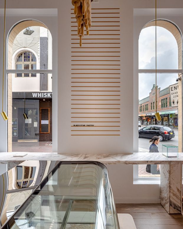 A refined commercial interior with a sculptural staircase or escalator descending through the foreground. Tall arched windows frame an urban streetscape, while the central wall features evenly spaced timber battens and a suspended sculptural timber light or art installation. A polished pale stone counter or ledge and reflective surfaces create a bright, gallery-like atmosphere.