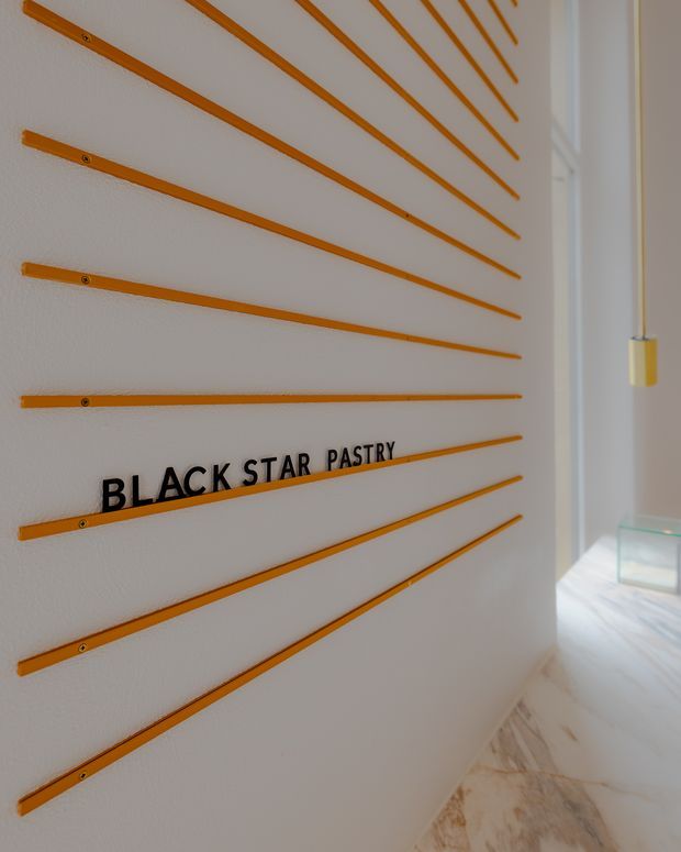 Interior view of a refined commercial pastry shop or hospitality space, featuring a white textured wall with radiating thin orange timber strips and raised black “BLACK STAR PASTRY” lettering. A slim pendant light, glass display case, bright glazing, and pale marble-like flooring are visible to the right.