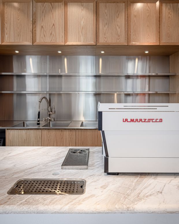 Commercial-style coffee bar or café back-of-house workspace with extensive warm timber cabinetry, stainless-steel shelving and splashback, a sink with gooseneck tap, and a large white espresso machine set on a pale veined stone counter. Integrated task lighting illuminates the preparation area.