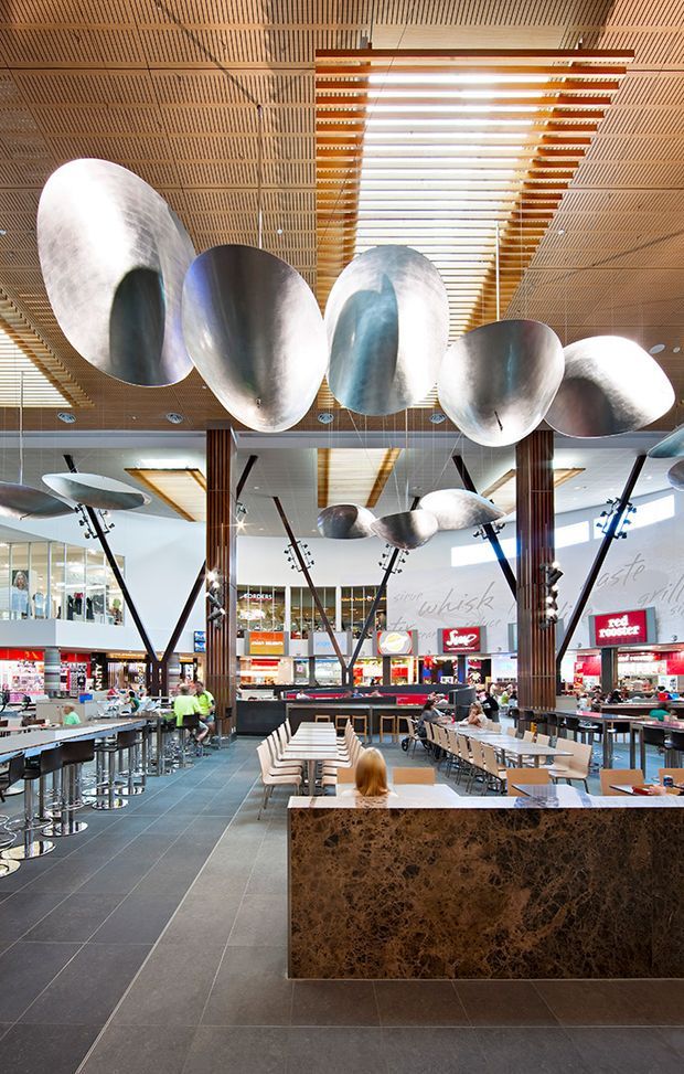 A spacious contemporary commercial food-court interior with large sculptural metallic pendant forms suspended beneath a slatted timber ceiling. The open dining area includes communal tables, bar-height seating, dark tiled flooring, timber-clad columns, and a prominent dark brown stone-clad counter. Retail and food outlets are visible in the background.