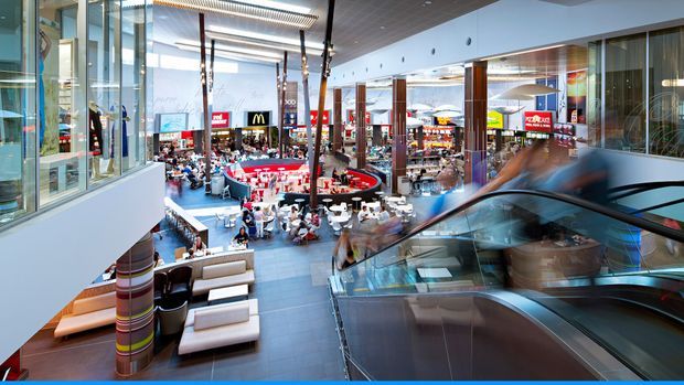 A bright, spacious multi-level indoor shopping centre atrium viewed from an escalator. The commercial space features extensive glazing, polished dark floor tiles, tall timber-clad structural columns, linear ceiling lighting, lounge seating, and a busy food-court area with visible retail signage.